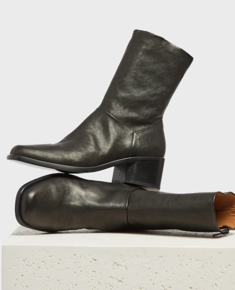 A pair of sleek, black leather ankle boots with a smooth texture, featuring a mid-height block heel, a square toe, and a soft tan interior lining, displayed on a textured white pedestal.