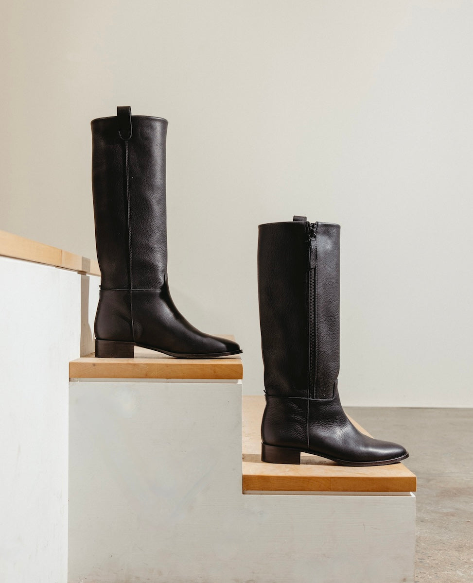 A pair of black knee-high leather boots is displayed on wooden steps against a minimalist background.